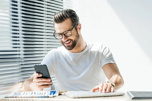 Hombre sonriente usando un teléfono inteligente mientras está sentado en el lugar de trabajo - foto de stock