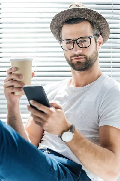 Handsome man in eyeglasses and hat holding coffee to go and using smartphone — Stock Photo