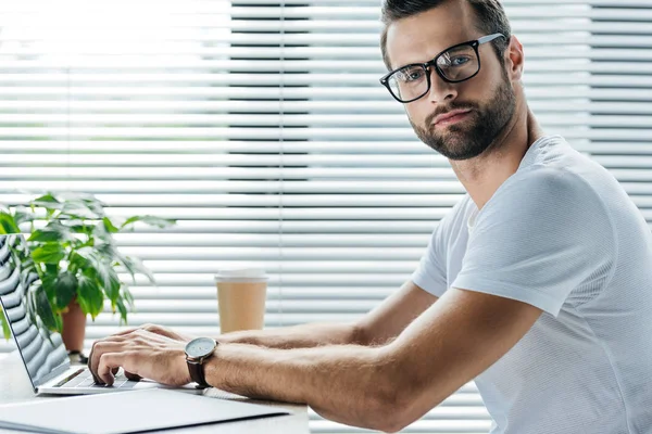 Hombre barbudo usando portátil mientras está sentado en el lugar de trabajo con una taza de café desechable — Stock Photo