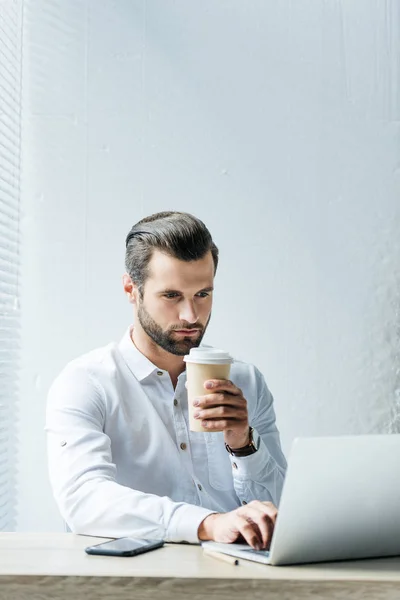Концентрированный бизнесмен держит кофе, чтобы пойти во время работы с ноутбуком — стоковое фото