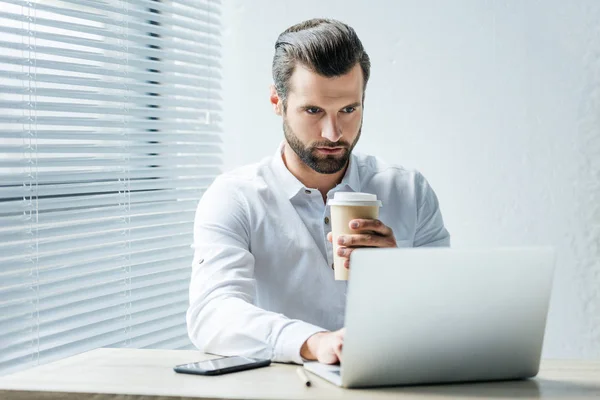 Guapo enfocado hombre de negocios sosteniendo café para ir mientras trabaja con el ordenador portátil en la oficina - foto de stock