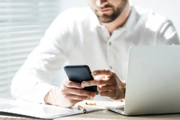 Cropped view of stylish businessman using smartphone at workplace with laptop and paperwork — Stock Photo