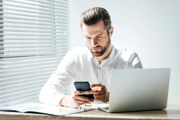 Homem de negócios elegante bonito usando smartphone no local de trabalho com laptop e documentos — Fotografia de Stock