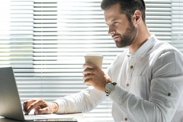 Концентрированный бизнесмен держит одноразовую чашку кофе и работает с ноутбуком у окна с жалюзи — стоковое фото