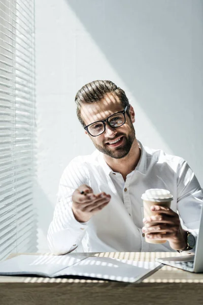Hombre de negocios sonriente en gafas de vista sentado en el lugar de trabajo con ordenador portátil y papeleo en pausa para el café - foto de stock