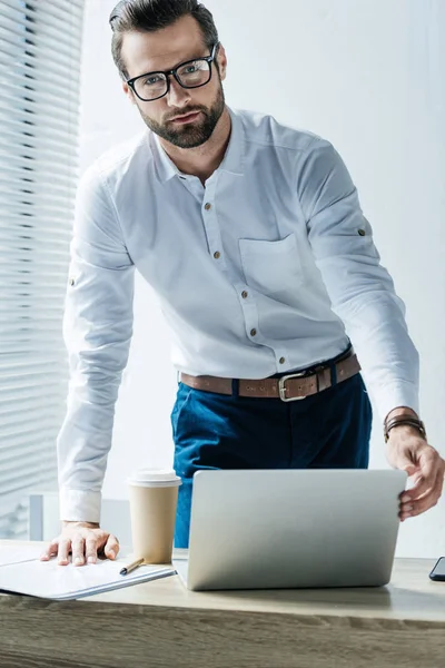 Красивый бородатый бизнесмен в офисе с ноутбуком и кофе, чтобы пойти — стоковое фото
