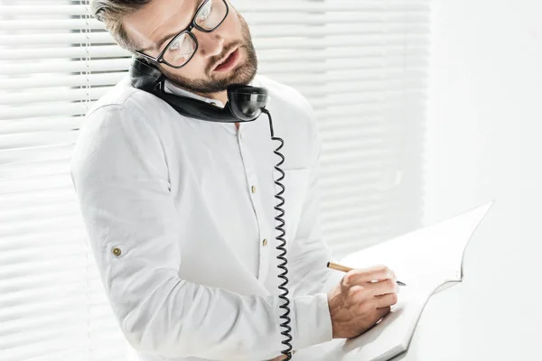 Bonito empresário falando no telefone rotativo enquanto escrevendo no notebook — Fotografia de Stock