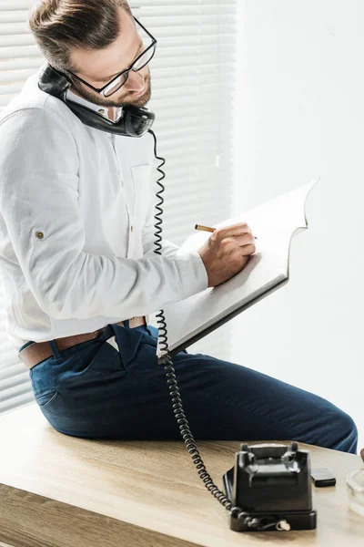 Empresário falando no telefone rotativo enquanto escreve no notebook — Fotografia de Stock