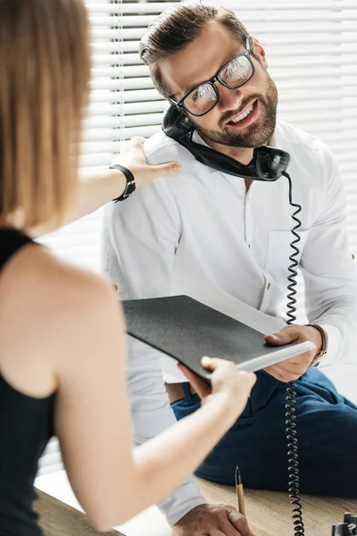Красивый улыбающийся бизнесмен разговаривает по ротационному телефону, в то время как секретарь дает ему папку — стоковое фото