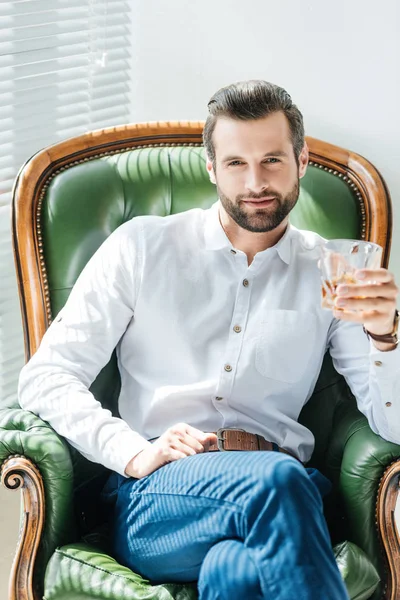 Hombre guapo sosteniendo vaso de whisky y sentado en un sillón verde cerca de la ventana - foto de stock