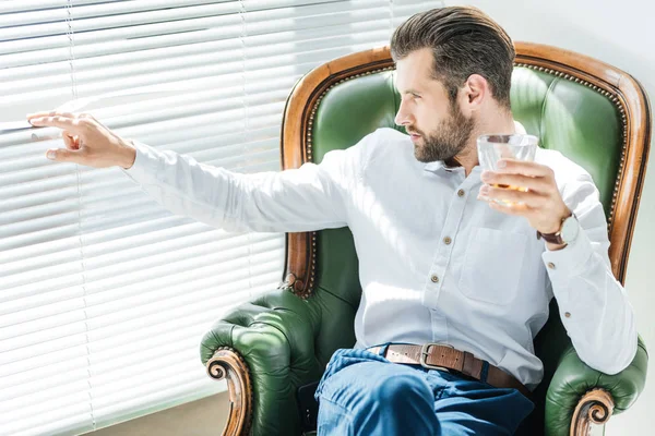 Bearded stylish man with glass of whiskey and sitting in green armchair and looking in window — Stock Photo