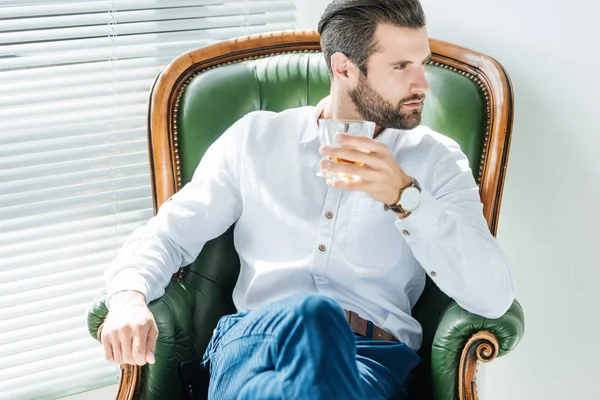 Elegante hombre de negocios elegante sosteniendo vaso de whisky y sentado en sillón - foto de stock