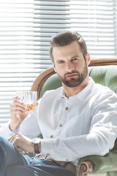 Confident stylish man holding glass of whiskey and sitting in armchair — Stock Photo
