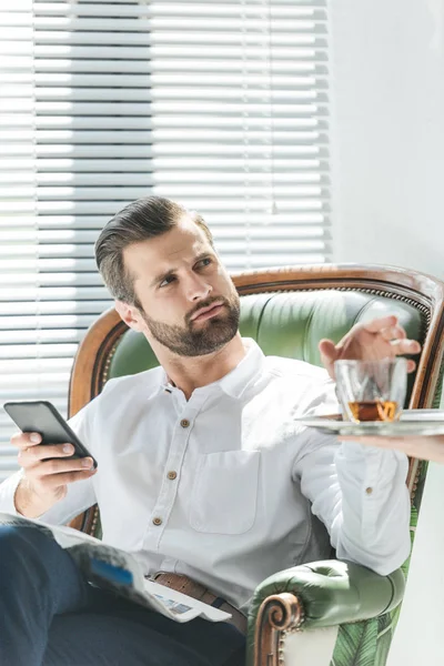 Hombre elegante guapo usando teléfono inteligente mientras toma el vaso de whisky de la bandeja - foto de stock