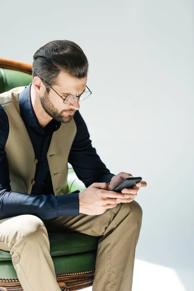 Élégant homme barbu dans des lunettes à l'aide d'un smartphone tout en étant assis dans un fauteuil vert — Photo de stock