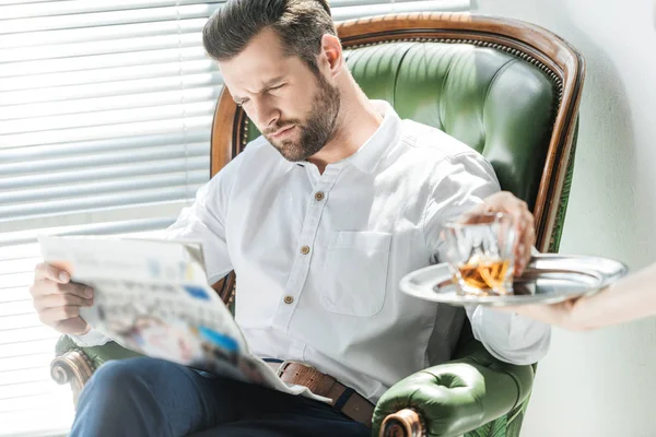 Stylish serious man reading newspaper while taking whiskey glass from tray — Stock Photo