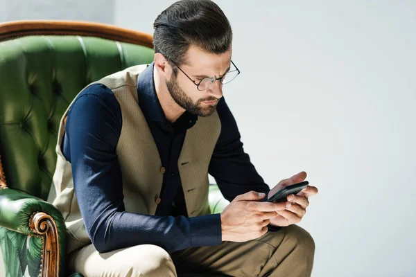 Homme barbu à la mode en utilisant smartphone tout en étant assis dans un fauteuil vert — Photo de stock