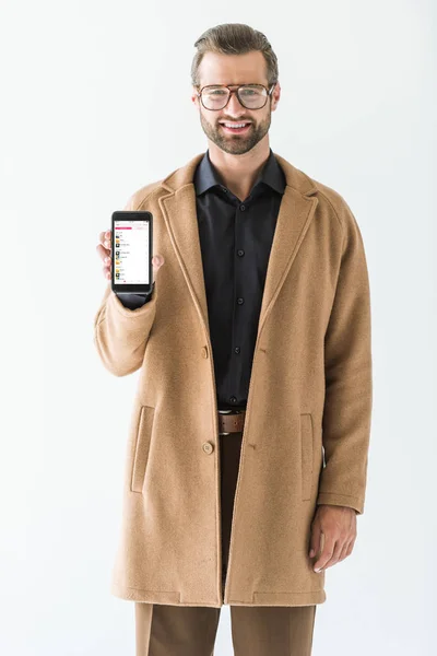 Hombre de moda en abrigo marrón presentando teléfono inteligente con aparato de música de manzana, aislado en blanco - foto de stock