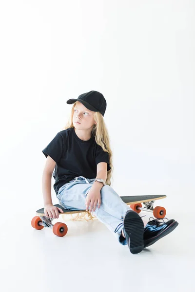 Entzückendes Kind mit langen lockigen Haaren, das auf einem Longboard sitzt und isoliert auf weißem Grund wegschaut — Stockfoto