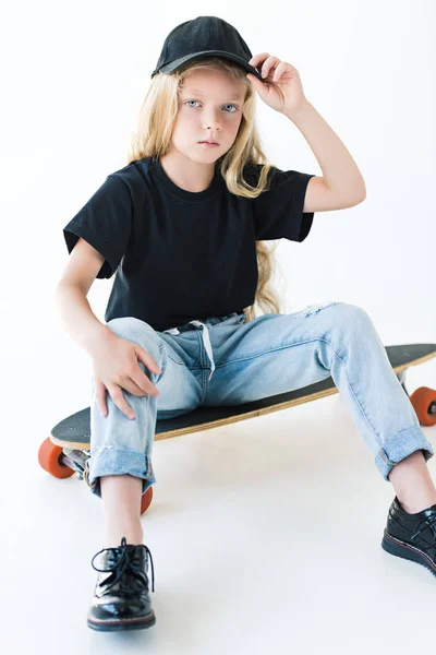 High angle view of kid in black cap and t-shirt sitting on longboard and looking at camera isolated on white — Stock Photo