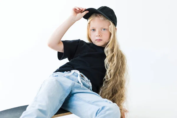 Schönes Kind mit langen lockigen Haaren, Schiebermütze und Blick in die Kamera, während es auf dem Skateboard sitzt, isoliert auf weiß — Stockfoto
