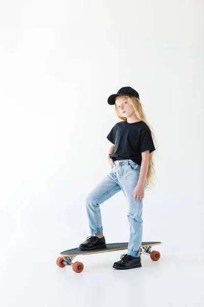 Full length view of cute kid in black cap and t-shirt standing on skateboard isolated on white — Stock Photo