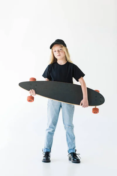 Lindo niño en gorra negra y camiseta sosteniendo longboard y mirando a la cámara aislada en blanco - foto de stock