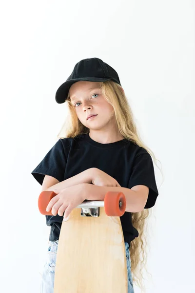 Hermoso niño en gorra negra y camiseta de pie con monopatín y mirando a la cámara aislada en blanco — Stock Photo
