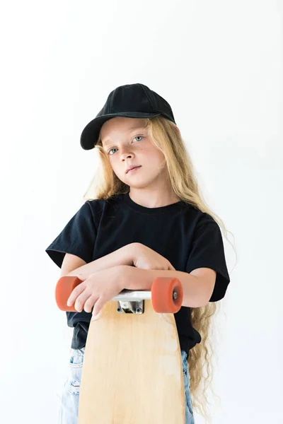 Hermoso niño en gorra negra y camiseta de pie con monopatín y mirando a la cámara aislada en blanco - foto de stock