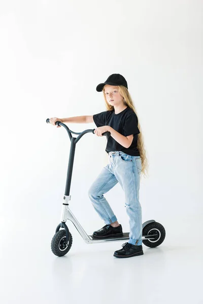 Cute stylish child in black t-shirt and cap riding scooter and looking at camera isolated on white — Stock Photo