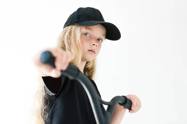 Beautiful kid in black t-shirt and cap riding scooter and looking at camera isolated on white — Stock Photo
