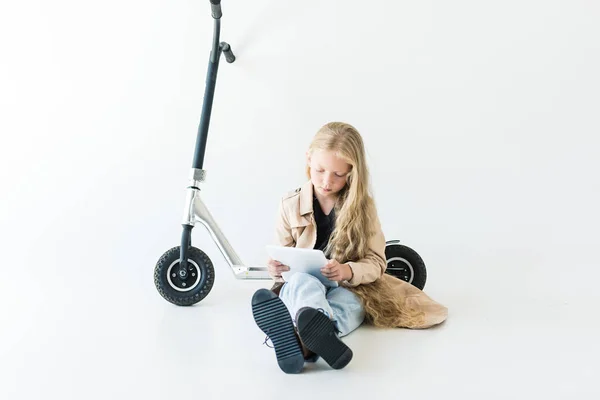 Hermoso niño con pelo rizado largo usando tableta digital mientras está sentado cerca de scooter aislado en blanco - foto de stock