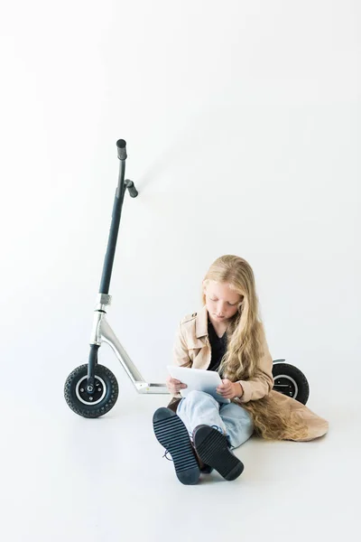 Очаровательный ребенок с длинными вьющимися волосами с помощью цифрового планшета, сидя рядом скутер изолирован на белом — стоковое фото