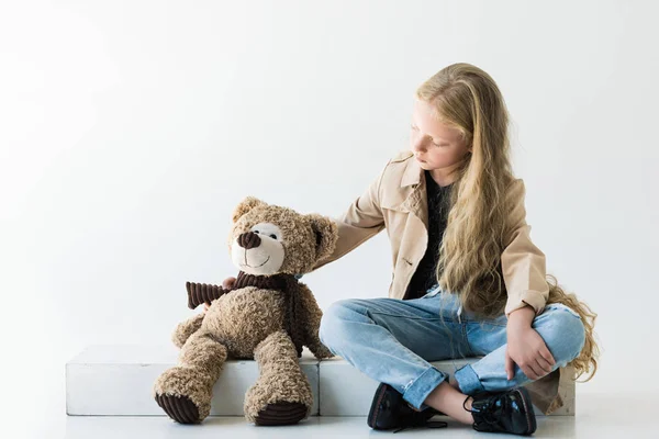 Vista de longitud completa de lindo niño elegante sentado y mirando oso de peluche en blanco - foto de stock