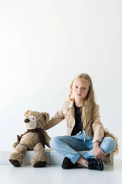 Belle enfant regardant la caméra tout en étant assis avec ours en peluche sur blanc — Photo de stock