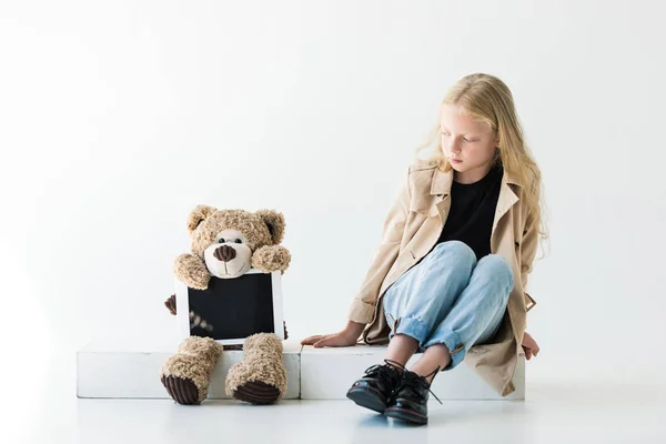 Schönes Kind mit langen lockigen Haaren schaut Teddybär mit digitalem Tablet auf weißem Hintergrund an — Stockfoto