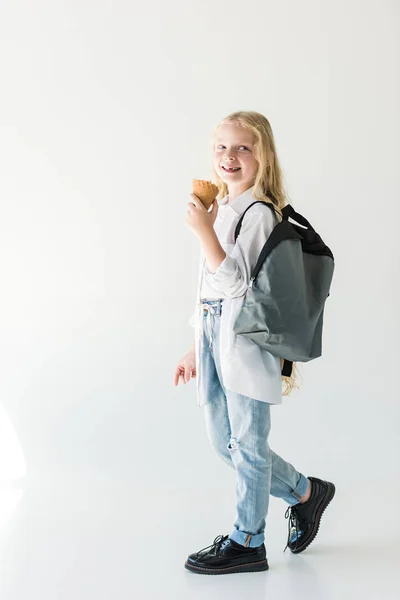 Vista completa de adorable niño feliz con mochila comiendo helado en blanco - foto de stock