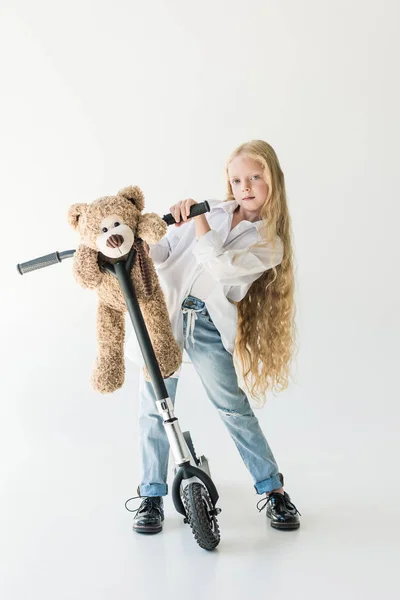 Adorable child with long curly hair standing with scooter and teddy bear, looking at camera on white — Stock Photo