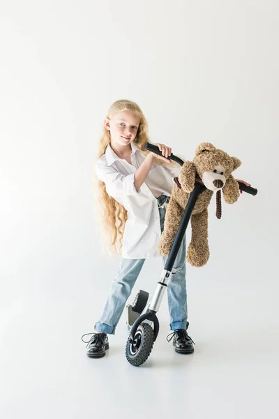 Adorable child with long curly hair standing with scooter and teddy bear, smiling at camera on white — Stock Photo