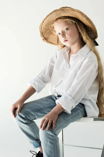 Hermoso niño en sombrero de mimbre sentado y mirando a la cámara en blanco - foto de stock