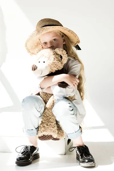 Vue pleine longueur de l'adorable enfant en chapeau de paille étreignant ours en peluche et regardant la caméra sur blanc — Photo de stock