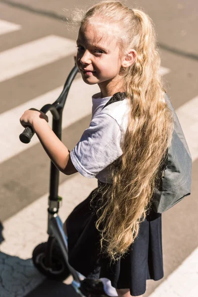 Vue grand angle de belle petite écolière avec de longs cheveux bouclés scooter d'équitation et regardant la caméra — Photo de stock