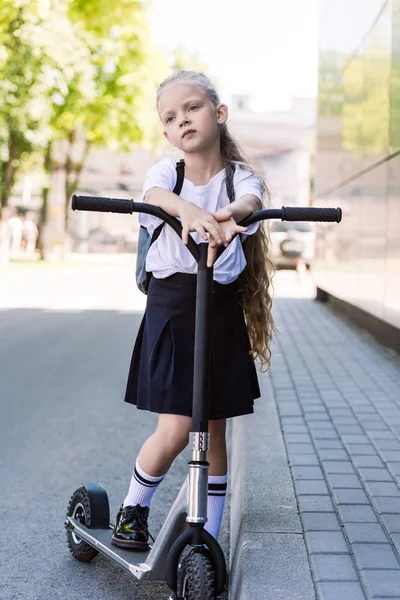 Mignon cher écolier avec sac à dos scooter d'équitation et regardant loin dans la rue — Photo de stock