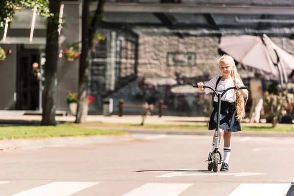 Beautiful little schoolgirl with long curly hair riding scooter at sunny day — Stock Photo