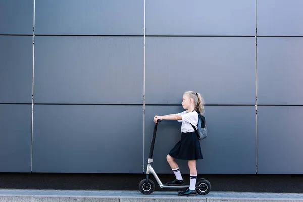 Vue latérale de adorable petite écolière avec de longs cheveux bouclés équitation scooter sur la rue — Photo de stock