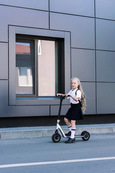Adorable little schoolgirl with long curly hair riding scooter and looking at camera on street — Stock Photo