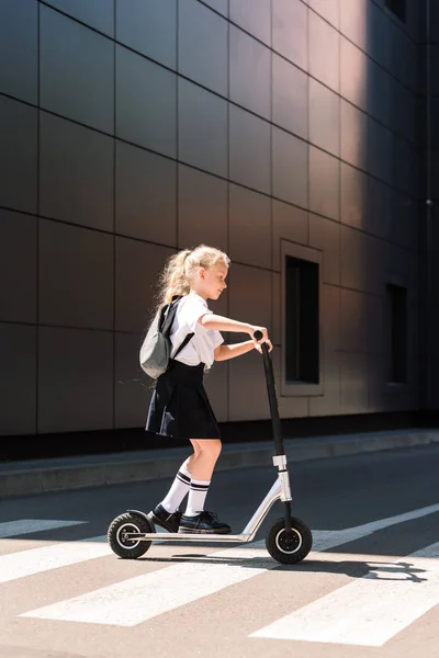 Side view of cute little schoolgirl with backpack riding scooter on street — Stock Photo