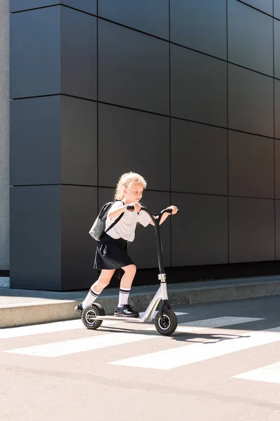 Cute little schoolgirl with backpack riding scooter on street — Stock Photo