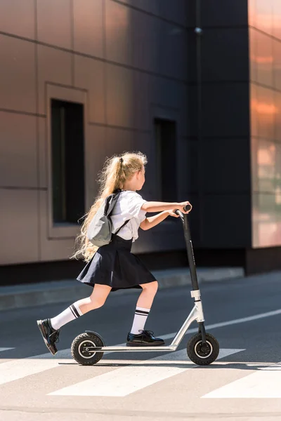 Вид сбоку симпатичной маленькой школьницы с рюкзаком, катающейся на скутере по улице — стоковое фото