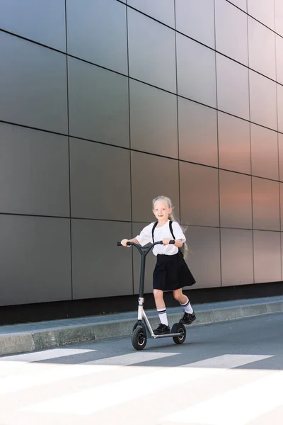 Vue pleine longueur d'adorable écolier équitation scooter et souriant à la caméra dans la rue — Photo de stock